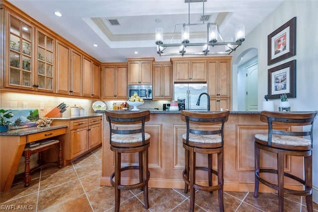 kitchen with appliances with stainless steel finishes, a kitchen breakfast bar, a raised ceiling, decorative light fixtures, and dark stone counters