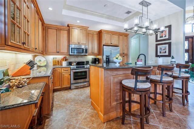 kitchen featuring stainless steel appliances, an island with sink, a raised ceiling, decorative light fixtures, and dark stone counters