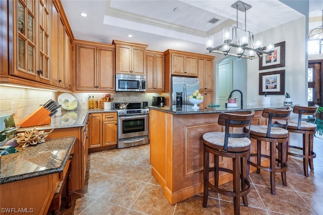 kitchen featuring decorative light fixtures, appliances with stainless steel finishes, a raised ceiling, an island with sink, and dark stone counters