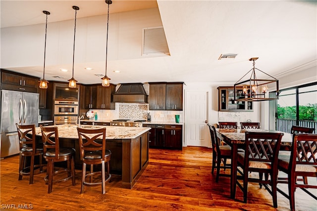 kitchen featuring decorative light fixtures, custom exhaust hood, appliances with stainless steel finishes, a kitchen island with sink, and an inviting chandelier