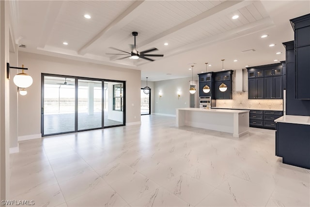 kitchen with wall chimney range hood, backsplash, ceiling fan, an island with sink, and beam ceiling