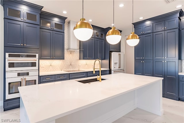 kitchen featuring wall chimney range hood, high end white fridge, an island with sink, stainless steel double oven, and tasteful backsplash