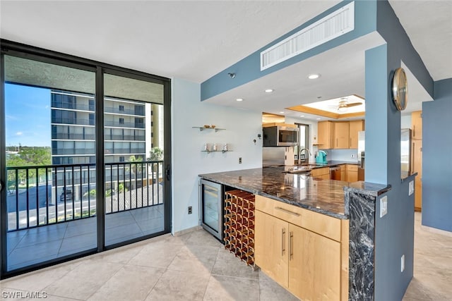 kitchen featuring dark stone counters, light brown cabinetry, kitchen peninsula, stainless steel appliances, and beverage cooler