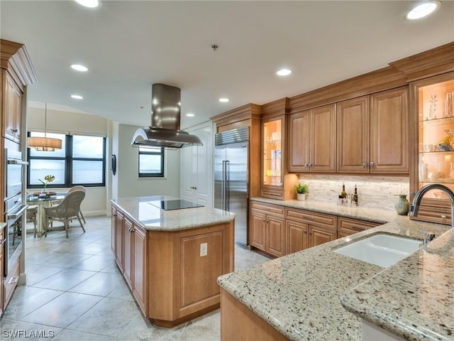 kitchen featuring built in fridge, island range hood, sink, light stone counters, and black electric cooktop