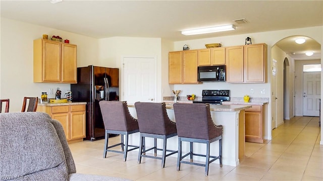 kitchen with light tile floors, a kitchen bar, light brown cabinetry, a center island with sink, and black appliances