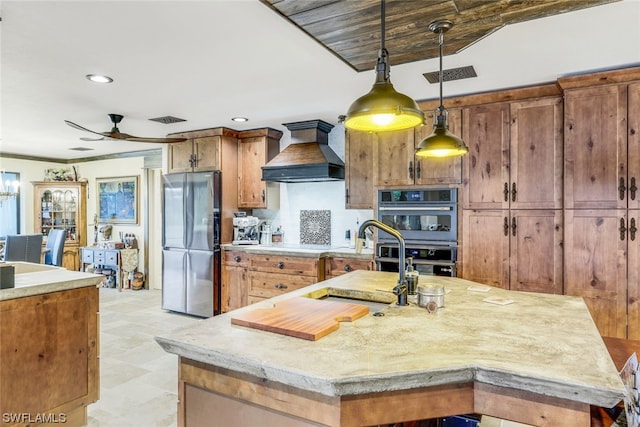 kitchen featuring ceiling fan, a center island with sink, stainless steel appliances, hanging light fixtures, and custom exhaust hood