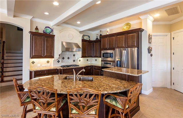 kitchen featuring appliances with stainless steel finishes, sink, an island with sink, wall chimney exhaust hood, and light stone counters