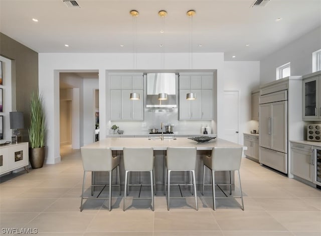 kitchen with a large island with sink, wall chimney range hood, paneled built in fridge, gray cabinets, and a breakfast bar