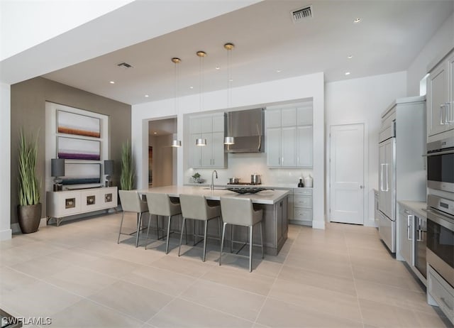 kitchen with an island with sink, a breakfast bar, wall chimney range hood, and gray cabinetry