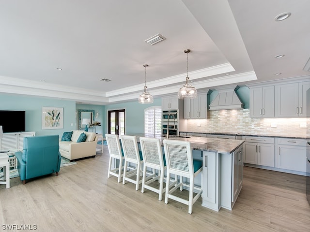 kitchen featuring a raised ceiling, light hardwood / wood-style flooring, custom range hood, a kitchen bar, and decorative light fixtures