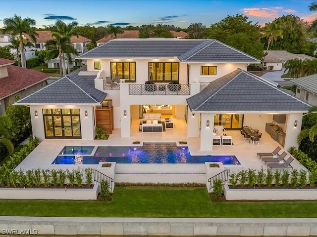 back house at dusk with an outdoor hangout area, a patio, a balcony, and a lawn