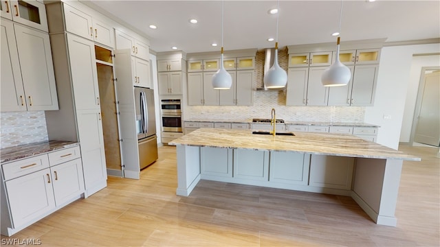 kitchen with an island with sink, stainless steel appliances, light stone countertops, wall chimney range hood, and tasteful backsplash