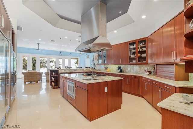 kitchen with light stone counters, a kitchen island with sink, island exhaust hood, ceiling fan, and tasteful backsplash