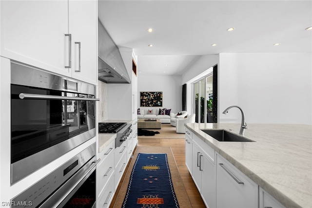 kitchen with wall chimney range hood, sink, light stone countertops, and white cabinets