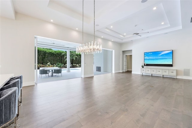 unfurnished living room with a raised ceiling, a high ceiling, ceiling fan with notable chandelier, and light hardwood / wood-style flooring