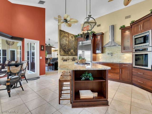 kitchen featuring backsplash, ceiling fan, a breakfast bar, wall chimney range hood, and built in appliances