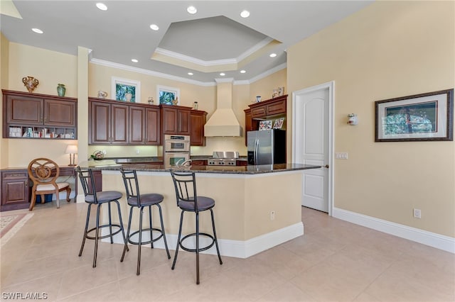 kitchen featuring custom exhaust hood, a large island with sink, appliances with stainless steel finishes, a raised ceiling, and a kitchen breakfast bar