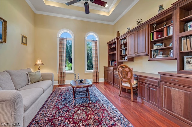 office area with a ceiling fan, a tray ceiling, dark wood finished floors, built in desk, and crown molding