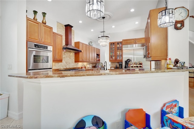 kitchen with kitchen peninsula, pendant lighting, light stone counters, appliances with stainless steel finishes, and wall chimney exhaust hood