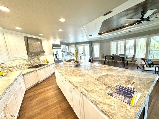 kitchen with light wood-type flooring, premium range hood, light stone counters, a tray ceiling, and stainless steel refrigerator with ice dispenser