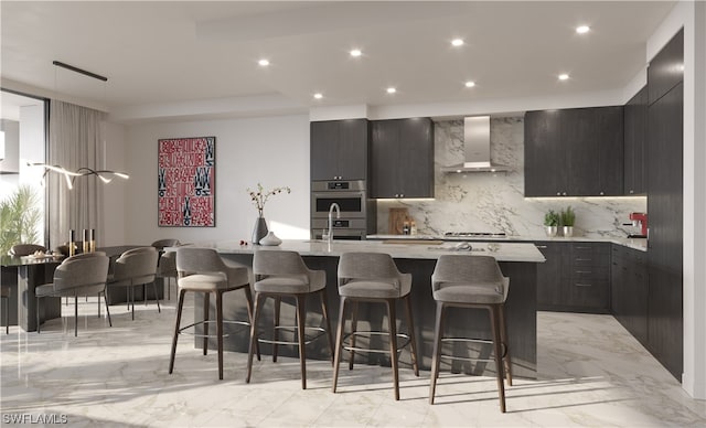 kitchen with an island with sink, double oven, light stone countertops, wall chimney exhaust hood, and backsplash