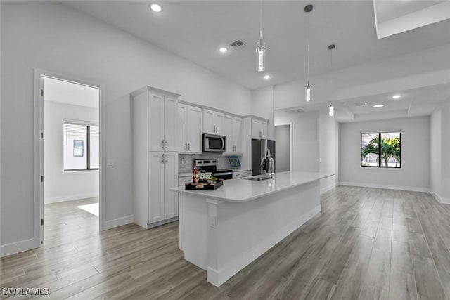 kitchen featuring appliances with stainless steel finishes, white cabinetry, hanging light fixtures, a center island with sink, and light hardwood / wood-style flooring