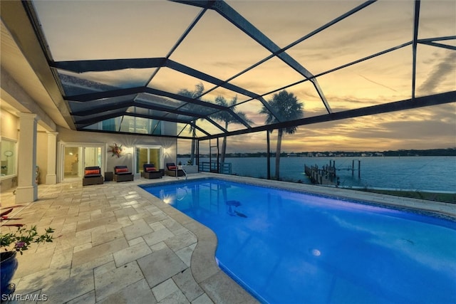 pool at dusk featuring a water view, a patio area, and glass enclosure