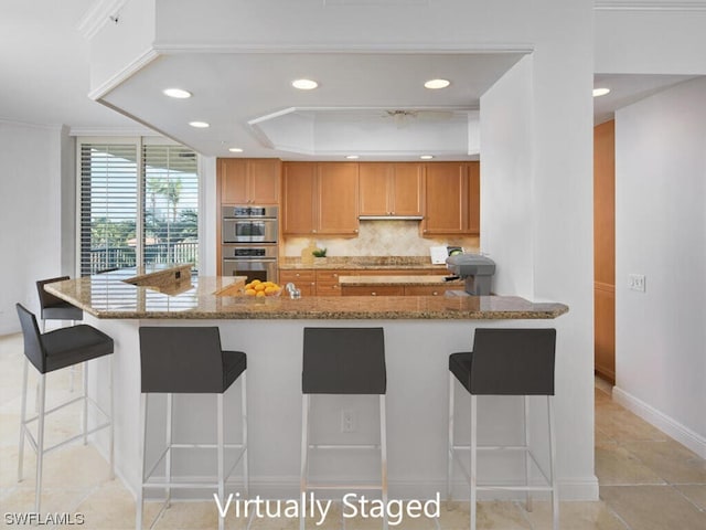 kitchen with backsplash, double oven, light tile flooring, a breakfast bar, and light stone counters