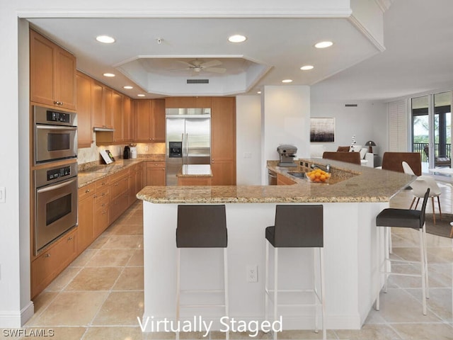 kitchen with a tray ceiling, ceiling fan, appliances with stainless steel finishes, light stone counters, and a breakfast bar area