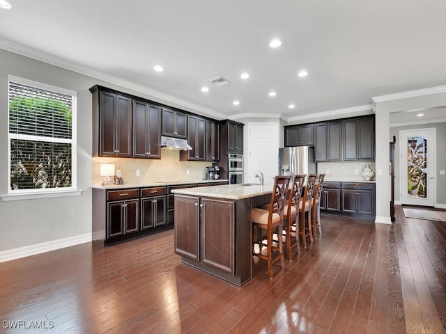 kitchen featuring appliances with stainless steel finishes, tasteful backsplash, ornamental molding, a kitchen island with sink, and dark brown cabinetry