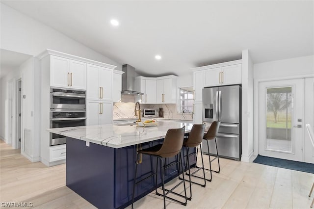 kitchen featuring a wealth of natural light, wall chimney exhaust hood, stainless steel appliances, a kitchen island with sink, and white cabinets