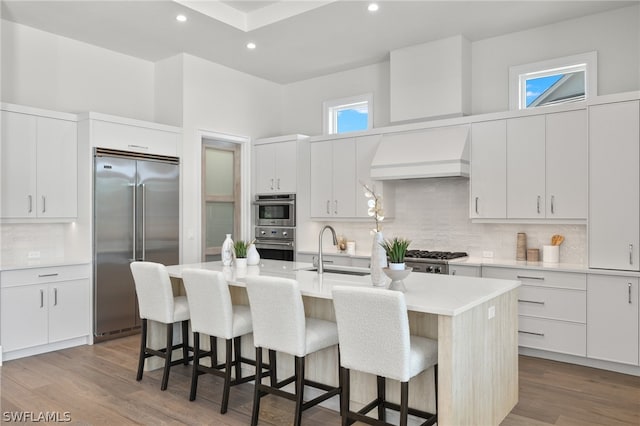 kitchen with custom range hood, a center island with sink, appliances with stainless steel finishes, backsplash, and wood-type flooring