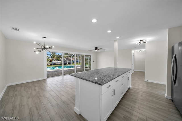 kitchen featuring stainless steel fridge, dark stone counters, a kitchen island, ceiling fan with notable chandelier, and white cabinetry