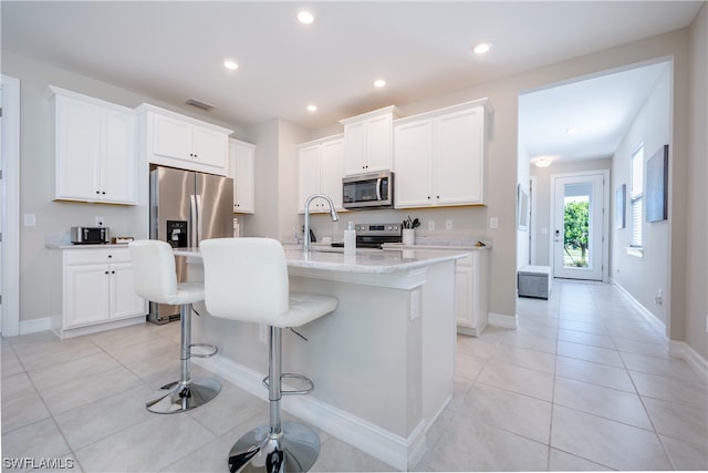 kitchen with light tile patterned flooring, white cabinetry, stainless steel appliances, light stone countertops, and a kitchen island with sink