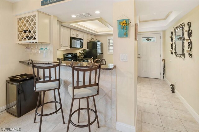 kitchen with a breakfast bar area, light tile patterned floors, black appliances, a raised ceiling, and cream cabinets