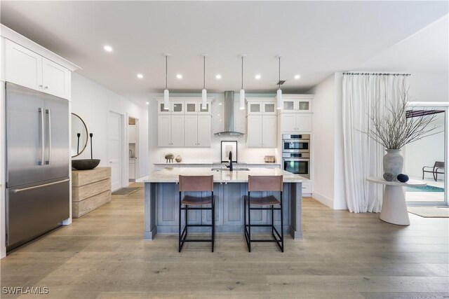 kitchen with a center island with sink, wall chimney range hood, white cabinetry, appliances with stainless steel finishes, and light wood-type flooring