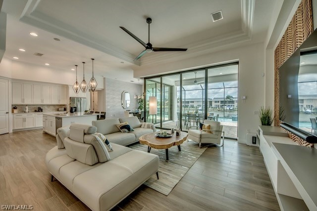 living room featuring ceiling fan with notable chandelier, a wealth of natural light, a tray ceiling, and light hardwood / wood-style flooring