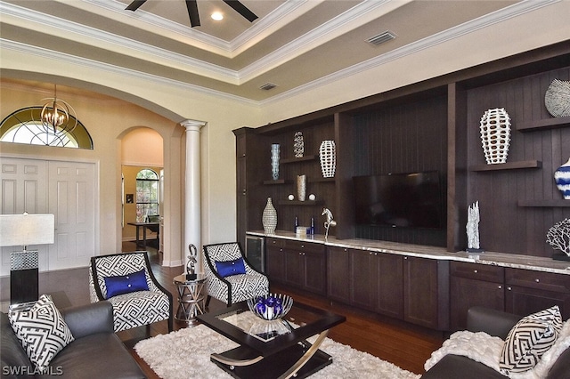 living room with a tray ceiling, dark wood-type flooring, ceiling fan with notable chandelier, decorative columns, and ornamental molding