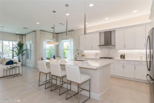 kitchen featuring hanging light fixtures, freestanding refrigerator, a kitchen island with sink, a sink, and wall chimney range hood