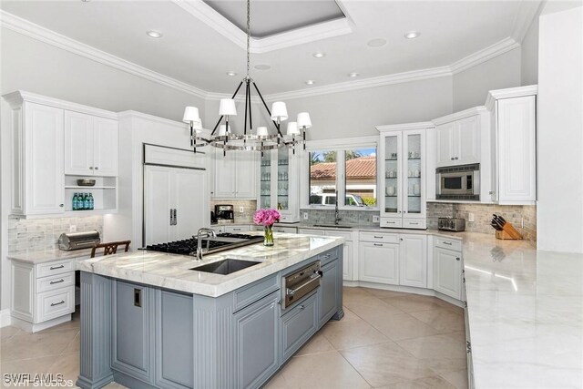 kitchen featuring built in appliances, light stone countertops, an island with sink, decorative light fixtures, and white cabinetry