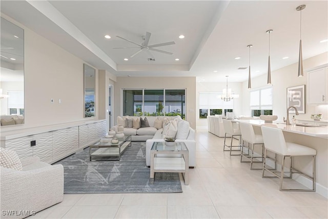 living room with light tile patterned floors, ceiling fan with notable chandelier, a tray ceiling, and sink