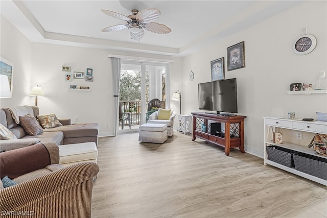 living area featuring a ceiling fan, a tray ceiling, baseboards, and light wood finished floors