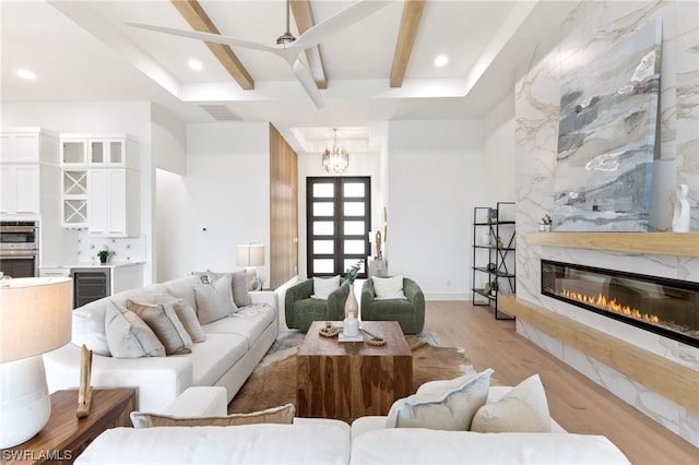 living room featuring wine cooler, beamed ceiling, ceiling fan with notable chandelier, a premium fireplace, and light hardwood / wood-style floors