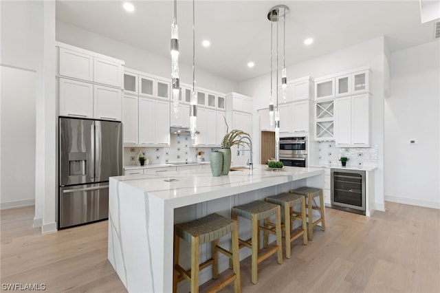 kitchen with beverage cooler, appliances with stainless steel finishes, light hardwood / wood-style floors, tasteful backsplash, and a center island with sink