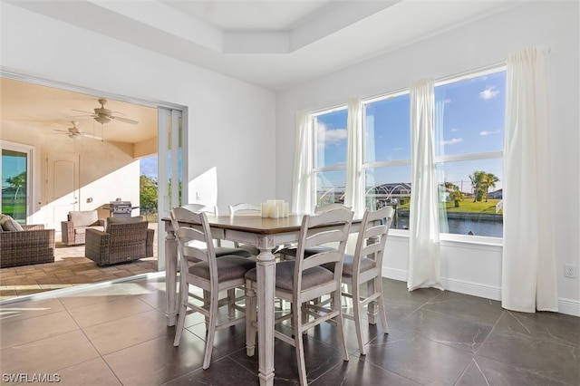 dining area with dark tile floors, ceiling fan, a water view, and a raised ceiling