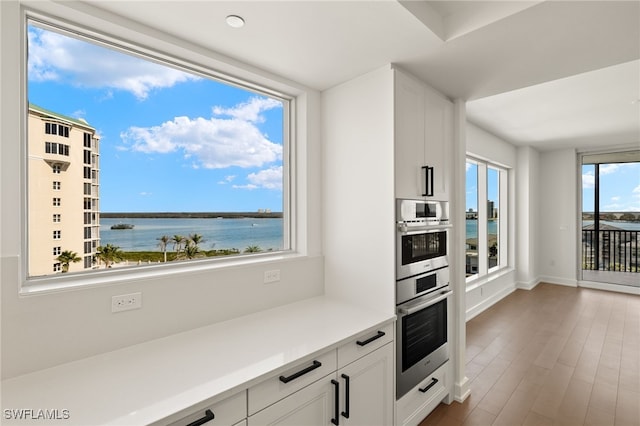 kitchen with light countertops, a water view, stainless steel double oven, white cabinets, and wood finished floors