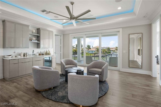 living room with ceiling fan, wine cooler, ornamental molding, wood-type flooring, and a tray ceiling