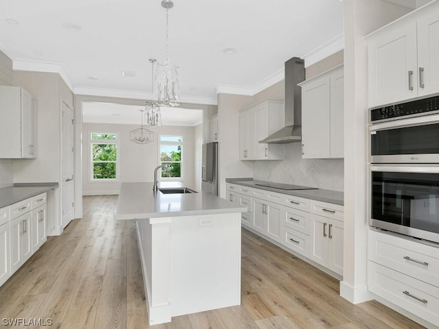 kitchen with an island with sink, stainless steel appliances, wall chimney range hood, white cabinetry, and sink