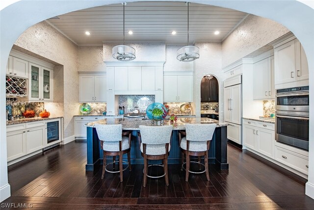 kitchen with an island with sink, light stone countertops, pendant lighting, stainless steel double oven, and dark hardwood / wood-style floors