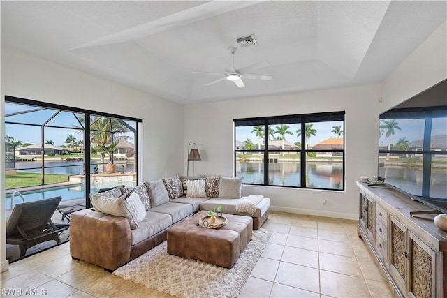 living room featuring ceiling fan, a raised ceiling, light tile flooring, and a water view